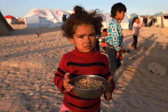 A young Palestinian girl holds a simple bowl of beans in a displacement camp in Gaza, a powerful human image of the humanitarian crisis unfolding under blockade and conflict.