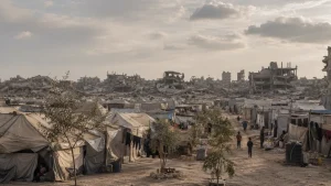 A refugee camp of beige tents and a dirt path in front of heavily damaged buildings and rubble under a cloudy sky.