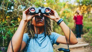 A woman uses binoculars while birdwatching on a wooded trail, with another hiker in the background.