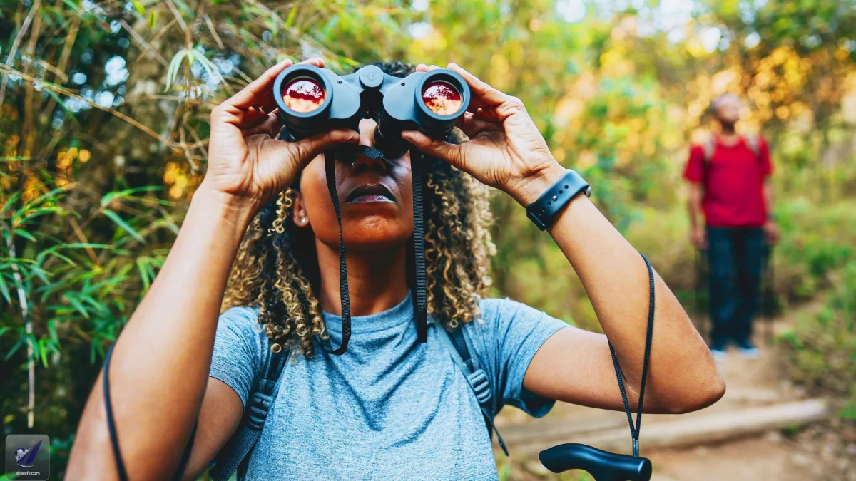 A woman uses binoculars while birdwatching on a wooded trail, with another hiker in the background.