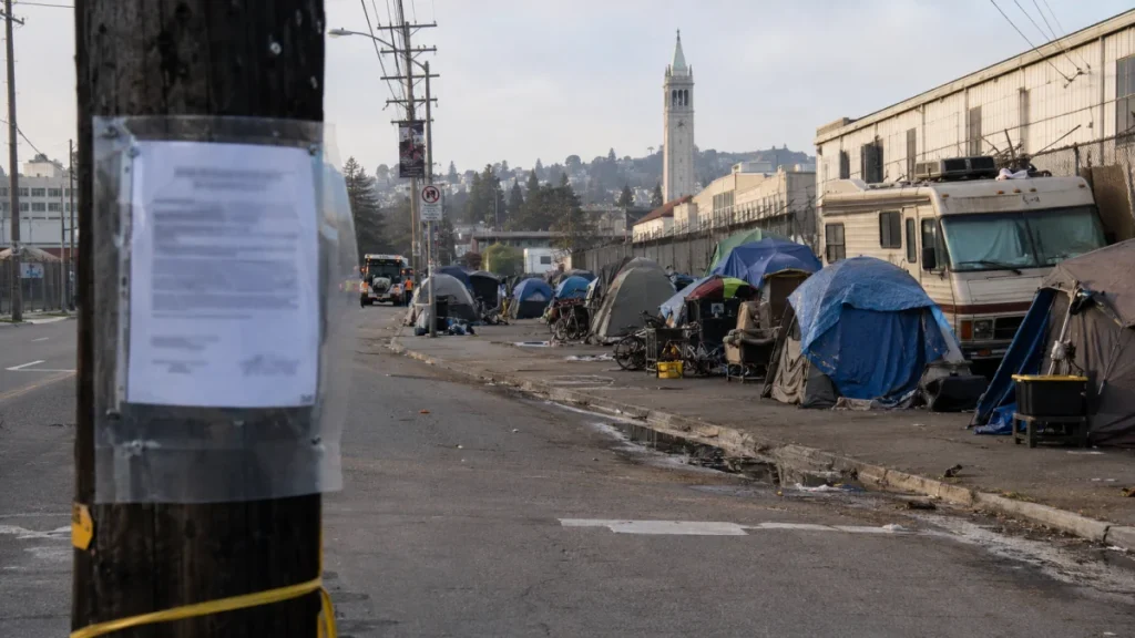 Tents and makeshift shelters line a Berkeley street beside parked RVs and a utility pole in the foreground.