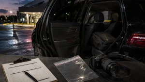Police or investigators near a black SUV with the rear door open, a clipboard and camera in the foreground, and a necklace visible on a bag inside the vehicle behind yellow caution tape at night.