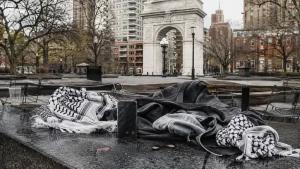A keffiyeh scarf, dark clothing, and a smartphone rest on a stone ledge in a city park with an arch monument in the background.