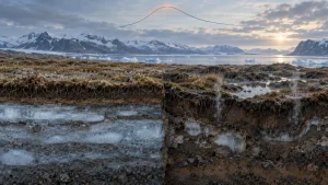 A frozen Arctic landscape with exposed permafrost layers cut into the ground beneath a snowy mountain range and cloudy sky.