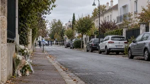 A quiet residential street with police tape in the distance and flowers left on the sidewalk near a wall.